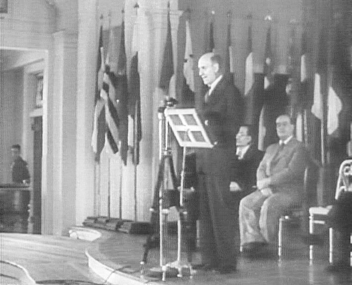 Photo d’archive en noir et blanc : un homme en costume parle au pupitre devant une rangée de drapeaux, à l’ouverture de la conférence de Bretton Woods en 1944.